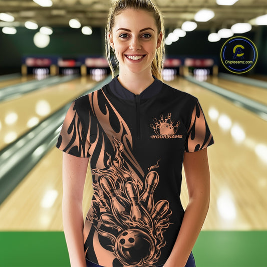 Female model front view inside bowling center wearing black quarter zip shirt with rose gold tribal flame bowling ball striking pins artwork