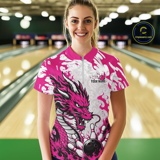 Person wearing a pink and white dragon-themed bowling shirt with a bowling ball graphic in a bowling alley.
