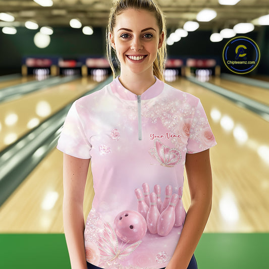 Female model shown from the front in a bowling alley wearing a quarter-zip shirt featuring glitter butterfly graphics, diamonds, and bowling artwork.