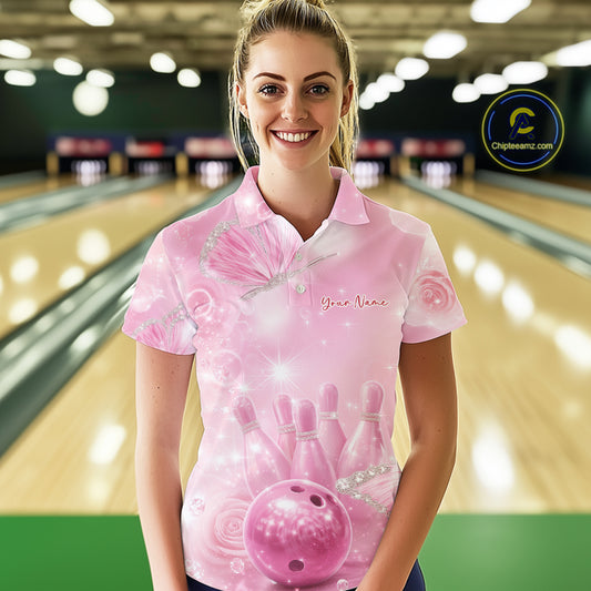 Female model at a bowling lane wearing a bowling polo shirt with pink glitter butterfly artwork, bowling pins, and a bowling ball on the front.