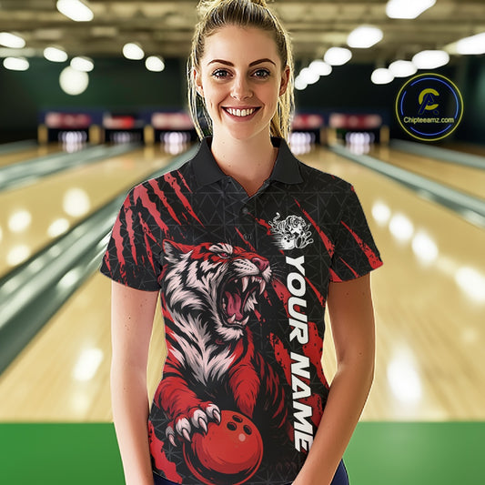 Front view of a female model at a bowling lane wearing a black and red bowling polo shirt showcasing a roaring tiger holding a red bowling ball with red claw-stripe graphics and a geometric background pattern.