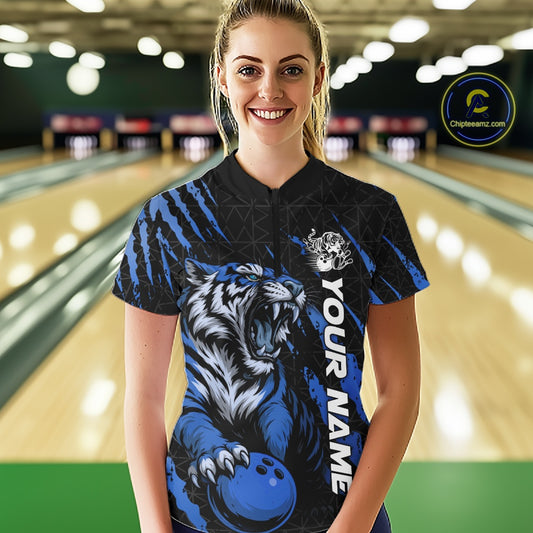 Front view of a female model at a bowling lane wearing a black and mid blue bowling quarter-zip shirt featuring a roaring tiger holding a mid blue bowling ball with coordinated claw-stripe graphics and a geometric background pattern.