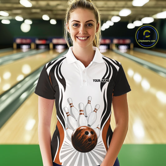 Female model at the bowling lane wearing a white, black, and orange bowling polo shirt, front view, featuring bowling ball striking pins graphics.