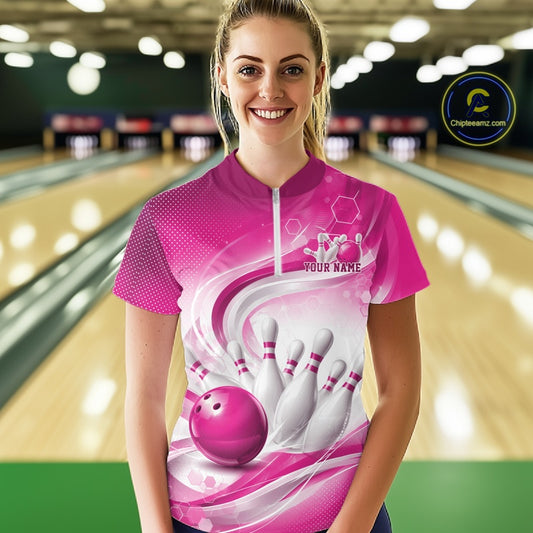 Female model standing in a bowling lane wearing a white and pink bowling quarter-zip shirt featuring abstract wave lines and a glossy bowling ball striking pins on the front.