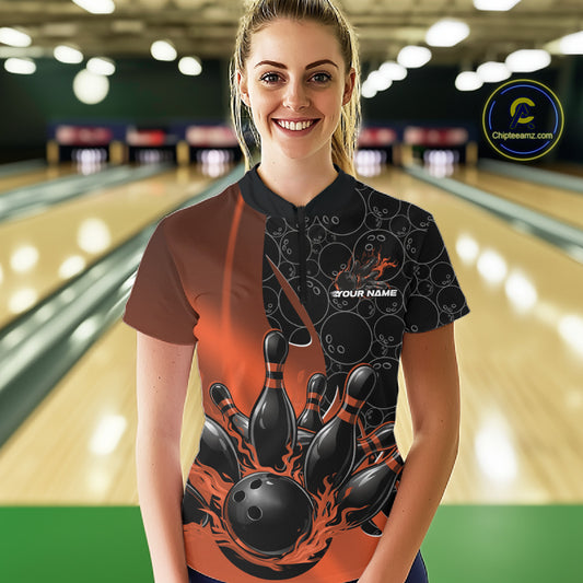 Female model in a bowling lane wearing black and orange quarter zip shirt, front view showcasing flaming bowling ball striking pins energetic design