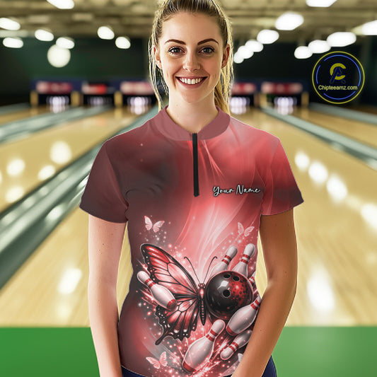 Female model wearing a red bowling quarter-zip shirt at a bowling lane with butterfly artwork, a bowling ball, and scattered bowling pins.