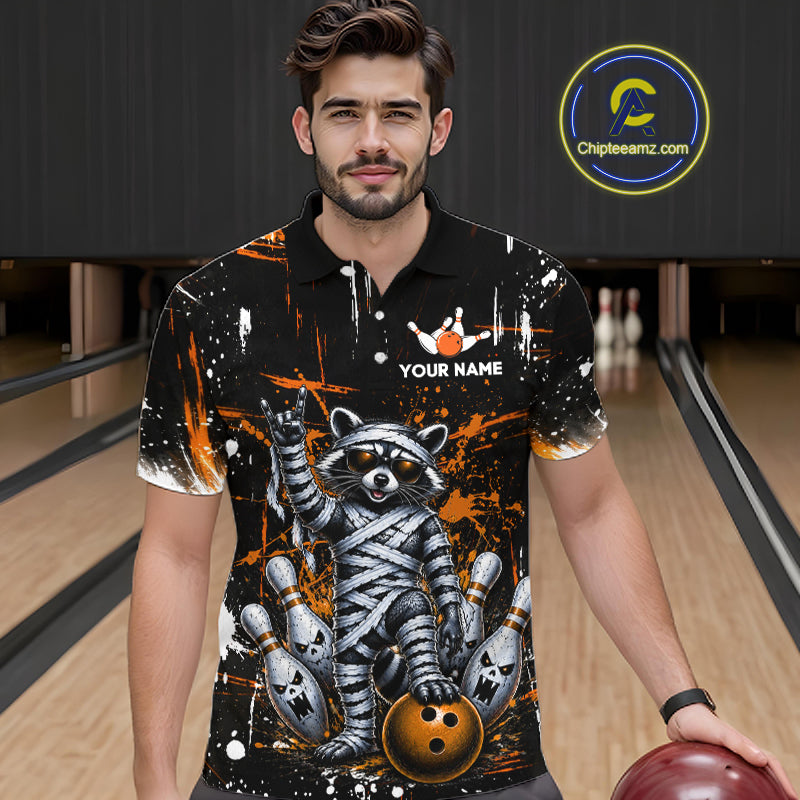 A man wearing a black short-sleeve bowling-themed polo shirt with a raccoon mummy graphic, orange paint splatters, and a white 'YOUR NAME' customization option, standing in a bowling alley.