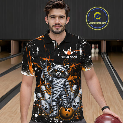 A man wearing a black short-sleeve bowling-themed polo shirt with a raccoon mummy graphic, orange paint splatters, and a white 'YOUR NAME' customization option, standing in a bowling alley.