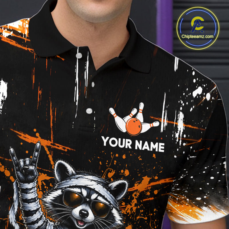 A close-up of a person wearing a black polo shirt with a racing raccoon graphic, orange paint splatters, white paint drips, and a bowling pin graphic above the text 'YOUR NAME'.