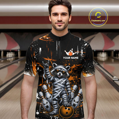 A man wearing a black short-sleeve bowling-themed quarter-zip shirt with a raccoon graphic, bowling pins, and orange paint splatter, set against a blurred bowling alley background. 