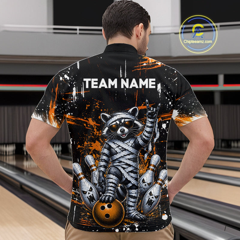A man viewed from behind wearing a black short-sleeve bowling shirt with a large graphic of a mummy raccoon holding bowling pins and a bowling ball. The shirt features the text 'TEAM NAME' on the back and is shown in a bowling alley setting.