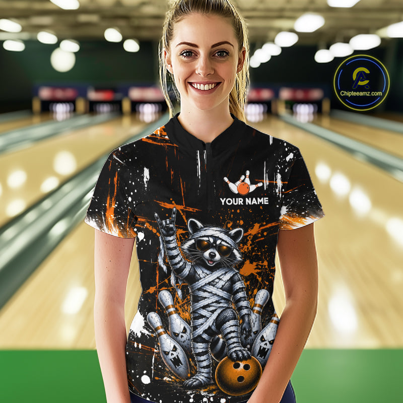 A woman standing in a bowling alley wearing a black short-sleeve bowling shirt with a raccoon mummy graphic and a white 'YOUR NAME' placeholder on the chest. 