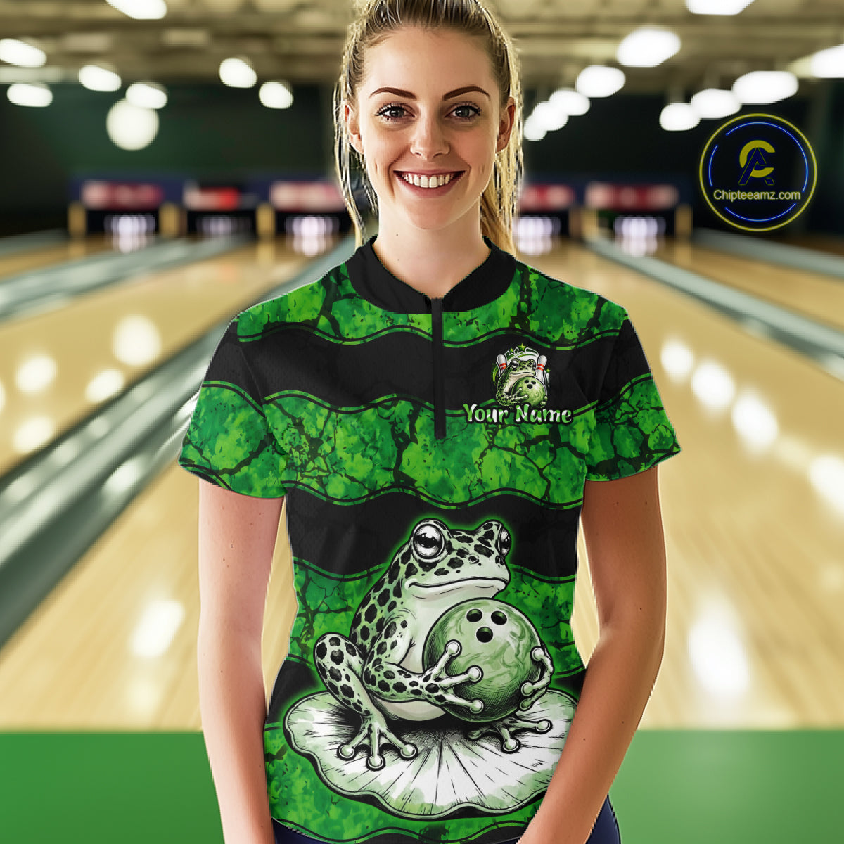 A woman stands in a bowling alley wearing a green and black short-sleeve bowling shirt with a frog and bowling ball graphic on the front. The shirt has a mock neck, black quarter-zip, and a custom name patch on the chest.