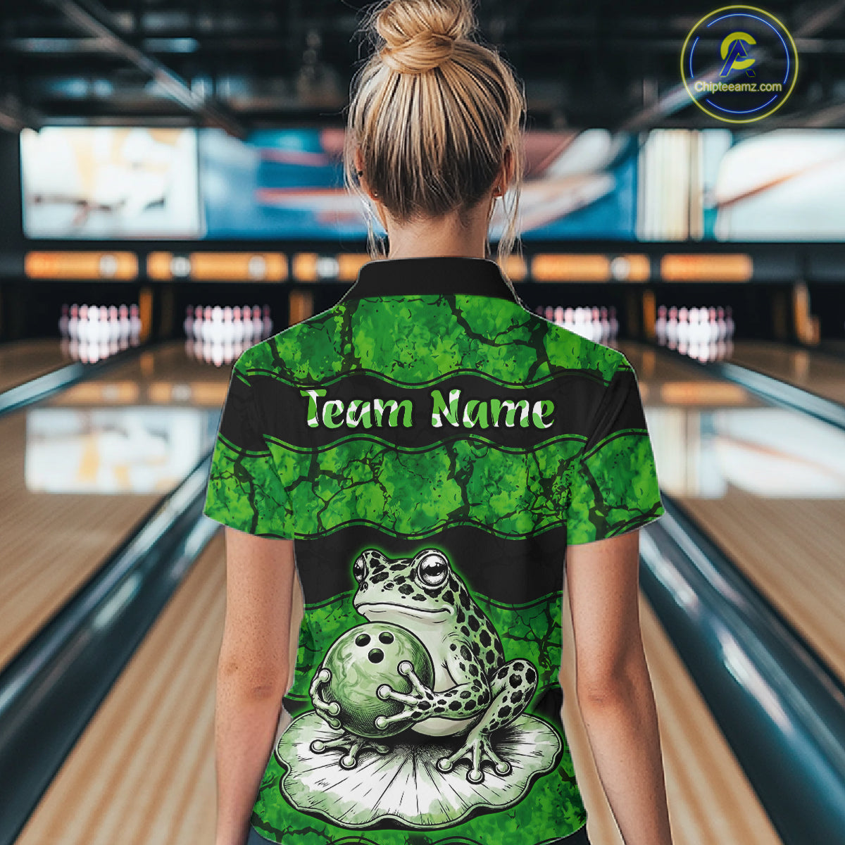 A woman viewed from behind wearing a green and black short-sleeve bowling shirt with a frog and bowling ball graphic on the back, standing in a bowling alley. The shirt has 'Team Name' printed across the upper back.