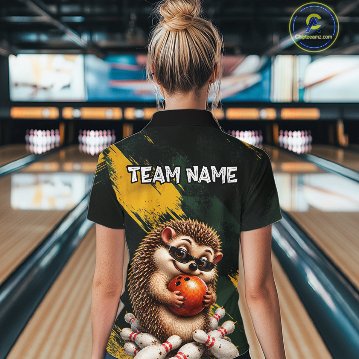 A woman viewed from behind wearing a black short-sleeve bowling-themed polo shirt with a large hedgehog graphic holding a bowling ball and the text 'TEAM NAME' on the back. The background shows blurred bowling lanes in an indoor bowling alley.