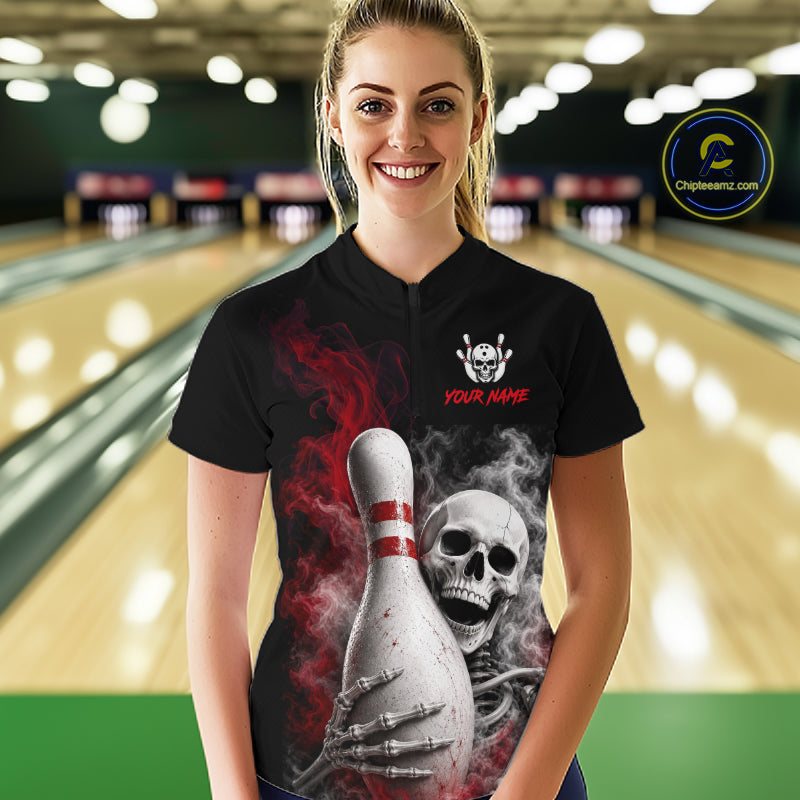 A woman stands in a bowling alley wearing a black short-sleeve bowling shirt with a large graphic print of a skull, skeleton hand, bowling pin, and red smoke effects. The shirt has a white skull-and-crossbones style logo on the chest with the text 'YOUR NAME'.