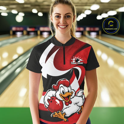 A woman standing in a bowling alley wearing a custom short-sleeve bowling shirt with a red, black, and white graphic design featuring a cartoon rooster holding a bowling ball. The shirt has a black collar and a short black zipper at the neck.