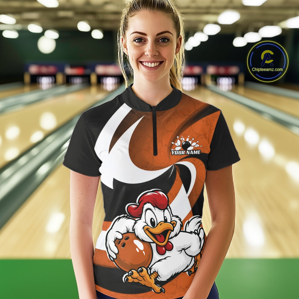 A woman standing in a bowling alley wearing a custom short-sleeve bowling shirt with a black and orange design, a white swoosh graphic, and a cartoon chicken holding a bowling ball. The shirt has a mock neck and a small 'YOUR NAME' placeholder on the chest.