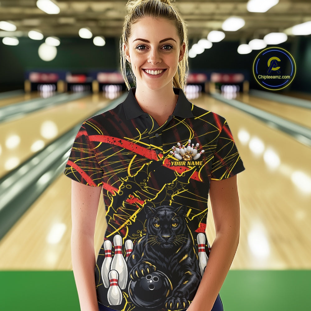A woman stands in a bowling alley wearing a black short-sleeve bowling t-shirt with a graphic print of a black panther, bowling pins, a bowling ball, and red and yellow abstract lines. The background shows blurred bowling lanes.
