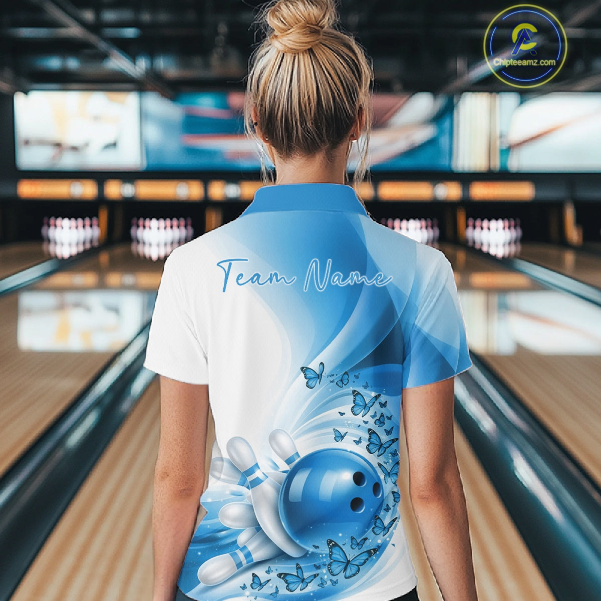 Back view of a women s white and blue bowling polo shirt displaying butterfly accents and bowling-themed graphics in a bowling venue.