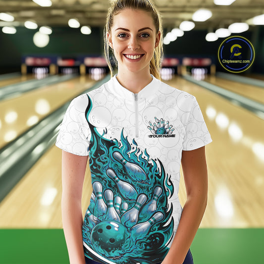 Front view female model at bowling lane wearing white and turquoise blue quarter zip shirt showing flaming bowling ball crashing into pins with vivid fire illustration.