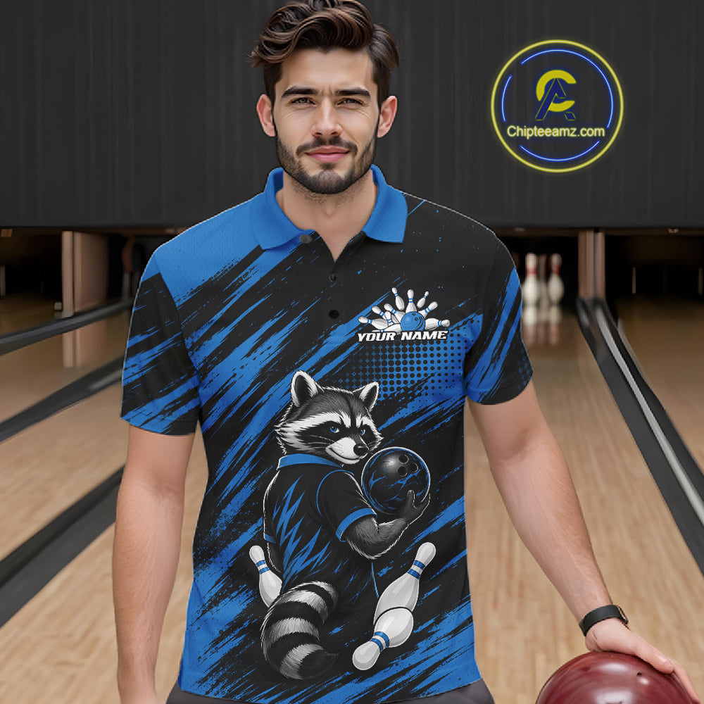 A man wearing a custom short-sleeve bowling polo shirt with a raccoon graphic, standing in a bowling alley. The shirt features a black and blue brush-stroke pattern with a raccoon holding a bowling ball and pins, and a customizable name print on the chest.