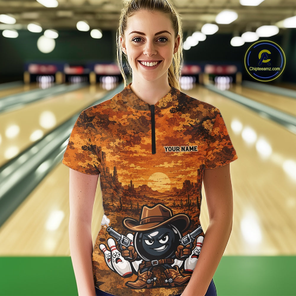 A woman stands in a bowling alley wearing a short-sleeve quarter-zip bowling shirt with a detailed western-themed graphic print featuring a cartoon bowling ball character. The shirt has a warm orange and brown sunset landscape design with the text 'YOUR NAME' on the chest.