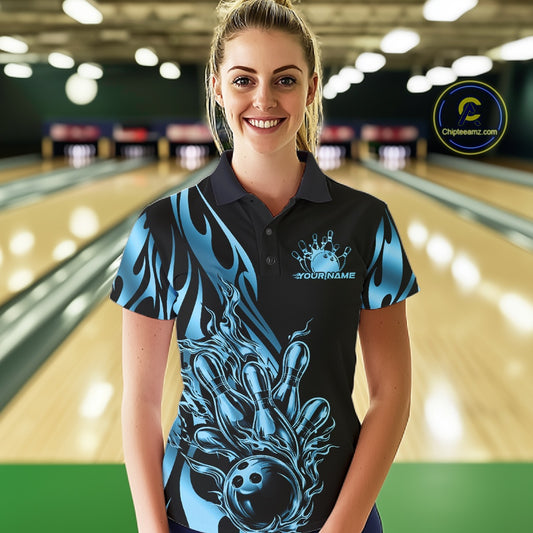 Female model seen from the front in a bowling lane wearing a black polo shirt featuring a blue gradient tribal flame design with a bowling ball striking pins in dynamic motion.