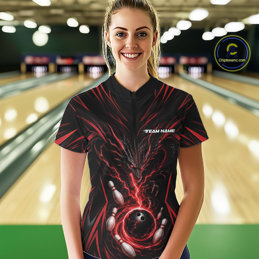 A woman wearing a black bowling-themed short-sleeve quarter zip shirt with red and black dragn and bowling graphics, standing in a bowling alley. 