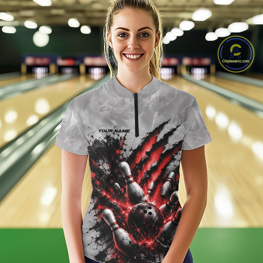 Front view of a female model wearing a gray bowling quarter zip shirt featuring a dramatic red bowling ball and pins graphic with claw-mark artwork in a bowling lane.