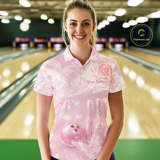Female model at a bowling lane wearing a bowling polo shirt with pink glitter butterfly artwork, roses, bowling pins, and a bowling ball on the front.