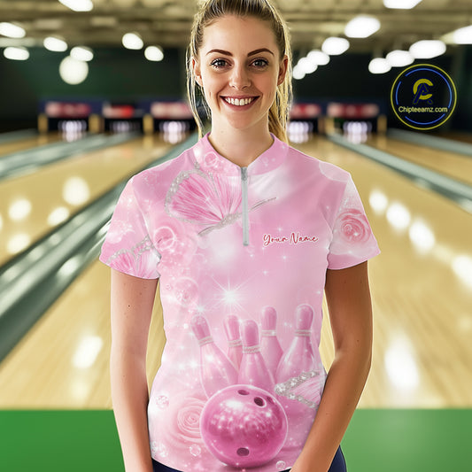 Female model at a bowling lane wearing a bowling quarter-zip shirt with pink glitter butterflies, bowling pins, and a bowling ball graphic on the front.