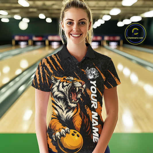 Front view of a female model at a bowling lane wearing a black and yellow bowling polo shirt featuring a roaring tiger holding a yellow bowling ball with orange claw-stripe graphics and a geometric background pattern.