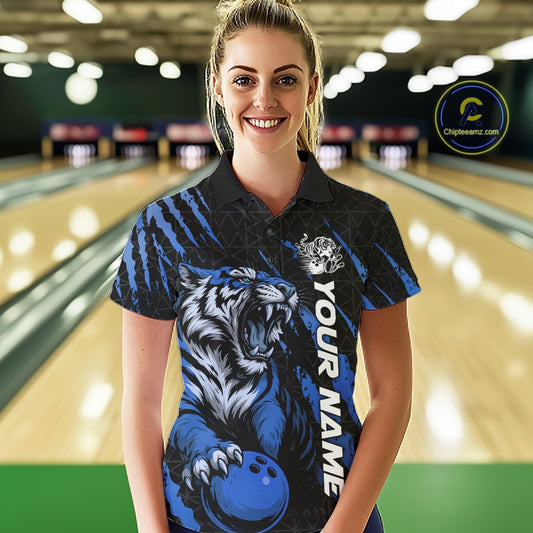 Front view of a female model at a bowling lane wearing a black and mid blue bowling polo shirt featuring a roaring tiger holding a mid blue bowling ball with coordinated claw-stripe graphics and a geometric background pattern.