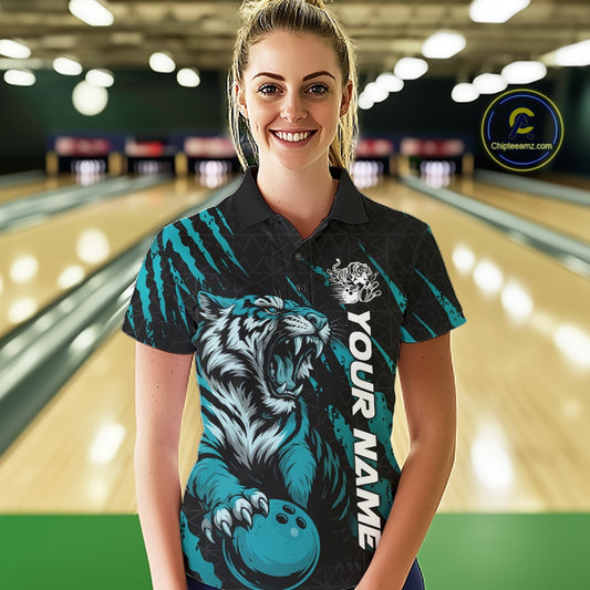 Front view of a female model at a bowling lane wearing a black and teal blue bowling polo shirt showcasing a roaring tiger holding a teal blue bowling ball with teal blue claw-stripe artwork and a geometric background pattern.