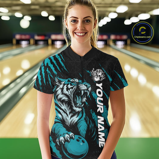 Front view of a female model at a bowling lane wearing a black and teal blue bowling quarter-zip shirt showcasing a roaring tiger holding a teal blue bowling ball with teal blue claw-stripe artwork and a geometric background pattern.