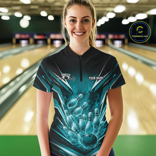 Front view female model at bowling lane wearing black and turquoise blue quarter zip shirt showing flaming bowling ball striking pins with energetic fire illustration.