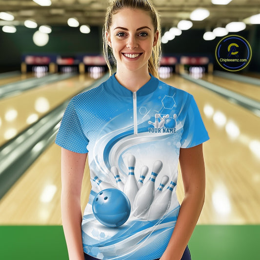Female model standing in a bowling lane wearing a white and blue bowling quarter-zip shirt featuring abstract wave lines and a glossy bowling ball striking pins on the front.