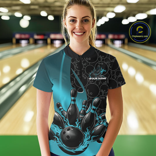 Front view of a female model wearing a black and blue bowling quarter-zip shirt at a bowling lane, featuring flaming bowling ball striking pins artwork with gradient ball pattern graphics.