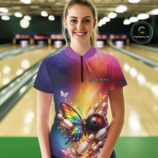 Female model at a bowling lane wearing a quarter-zip bowling shirt with a rainbow butterfly, bowling ball, and scattered pin artwork.